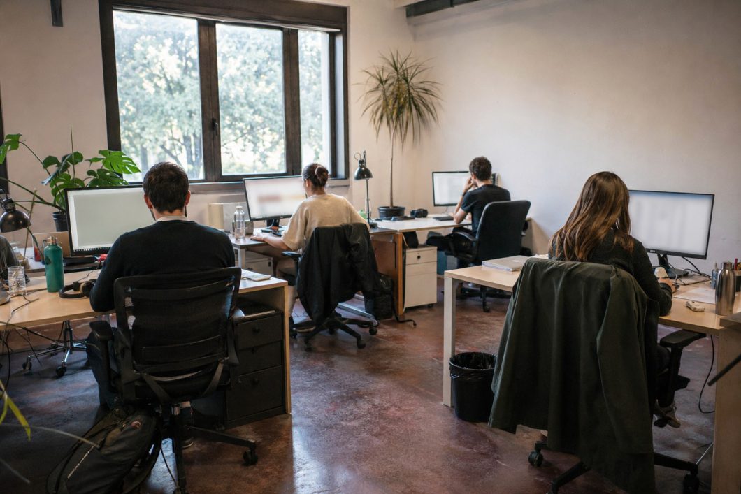 Quattro persone al lavoro alle proprie postazioni in un ufficio open space con piante e grandi finestre.